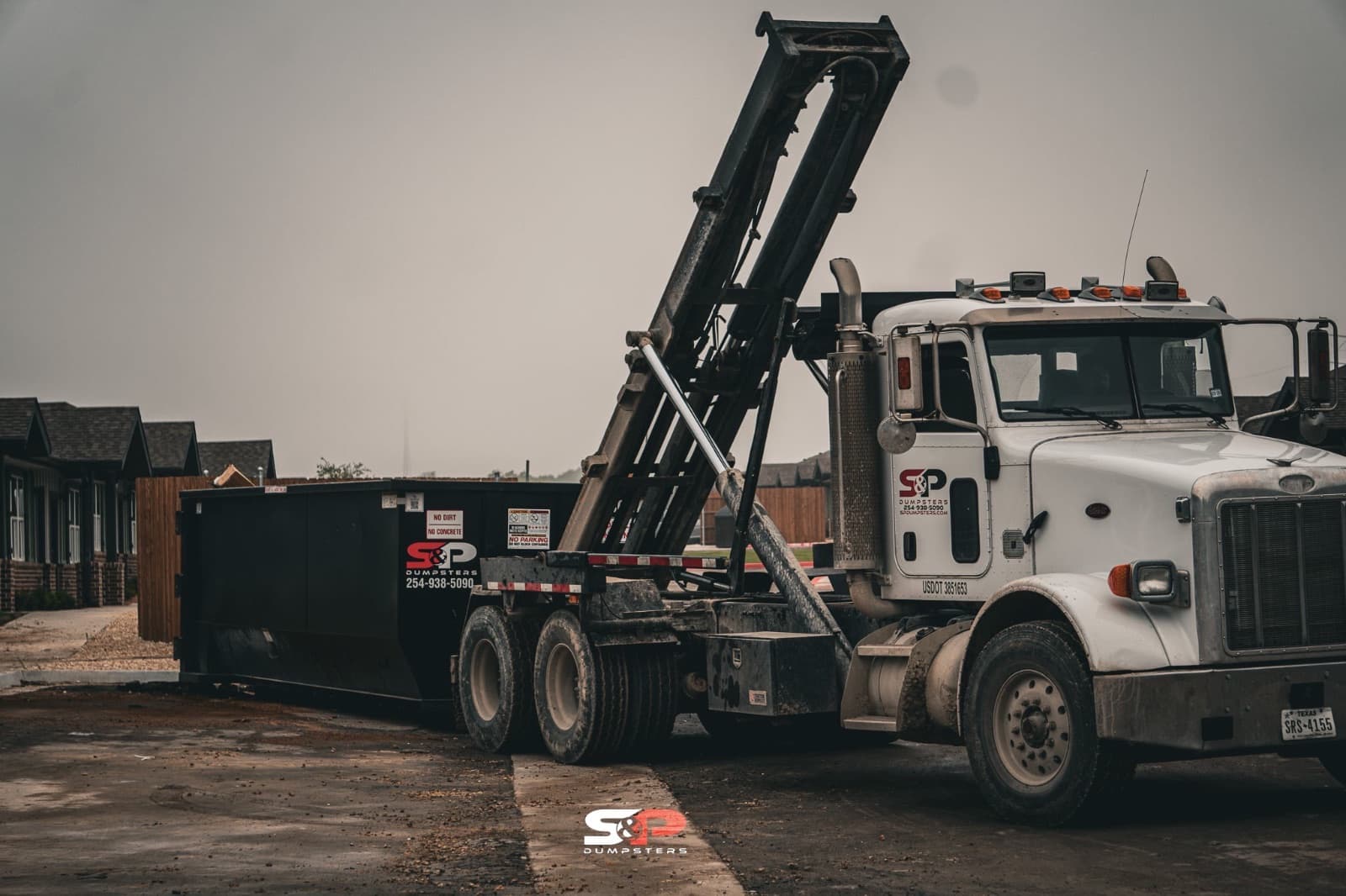 An S&P Dumpsters roll-off truck hauling a loaded container past new homes